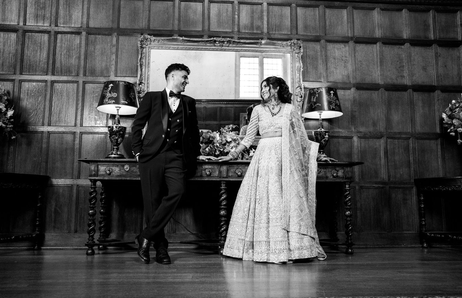 Black and white photo of an Indian couple in traditional wedding attire holding hands in an elegant wood-paneled ballroom.