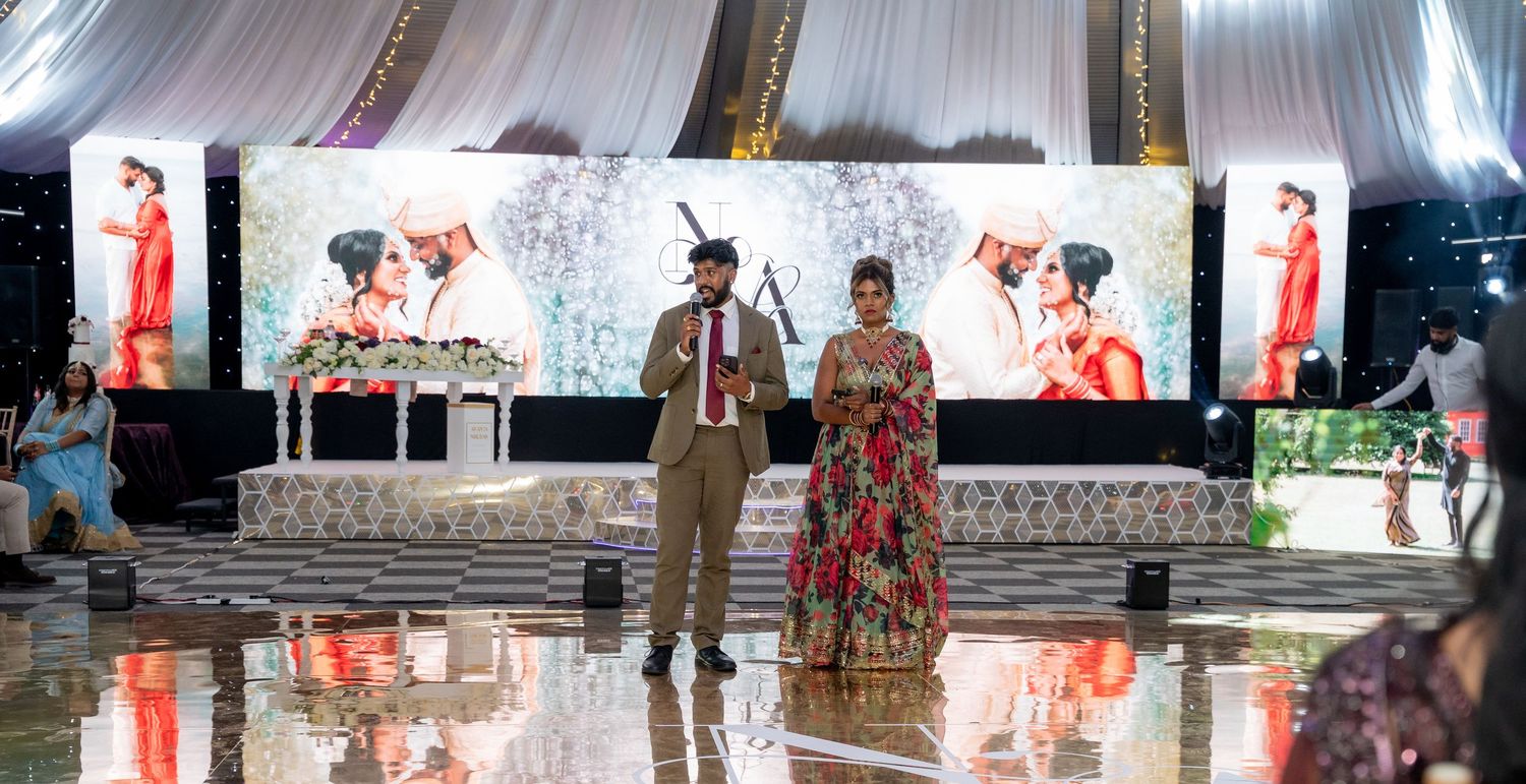 Couple in elegant attire speaking at a grand Indian wedding reception with large LED display backdrop.