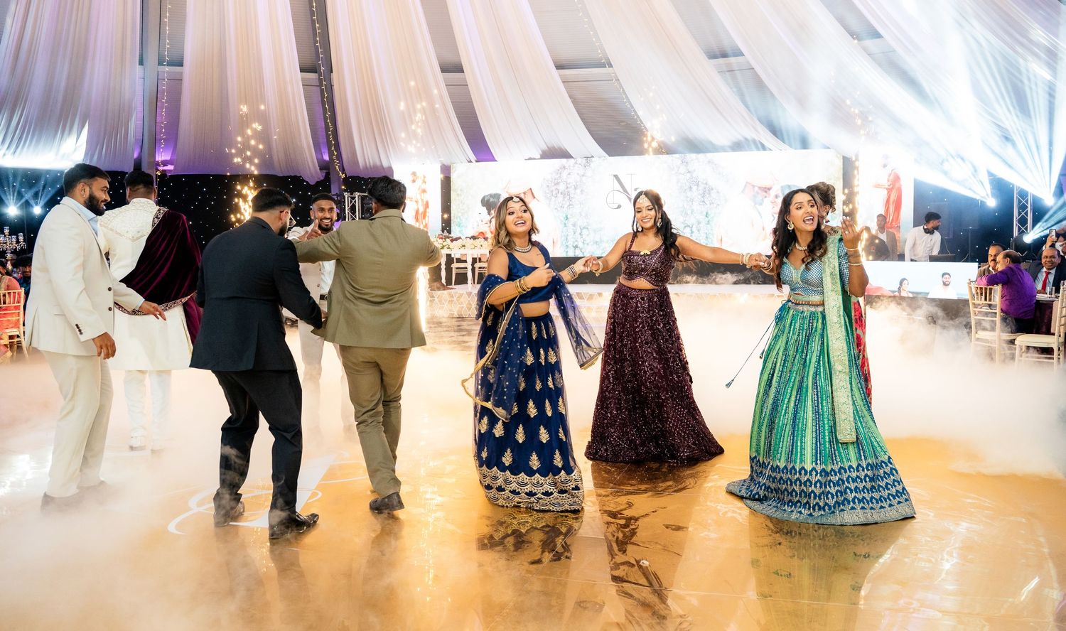 Guests in vibrant Indian attire dance joyfully at an elegant wedding reception with flowing white drapes and fog effects.