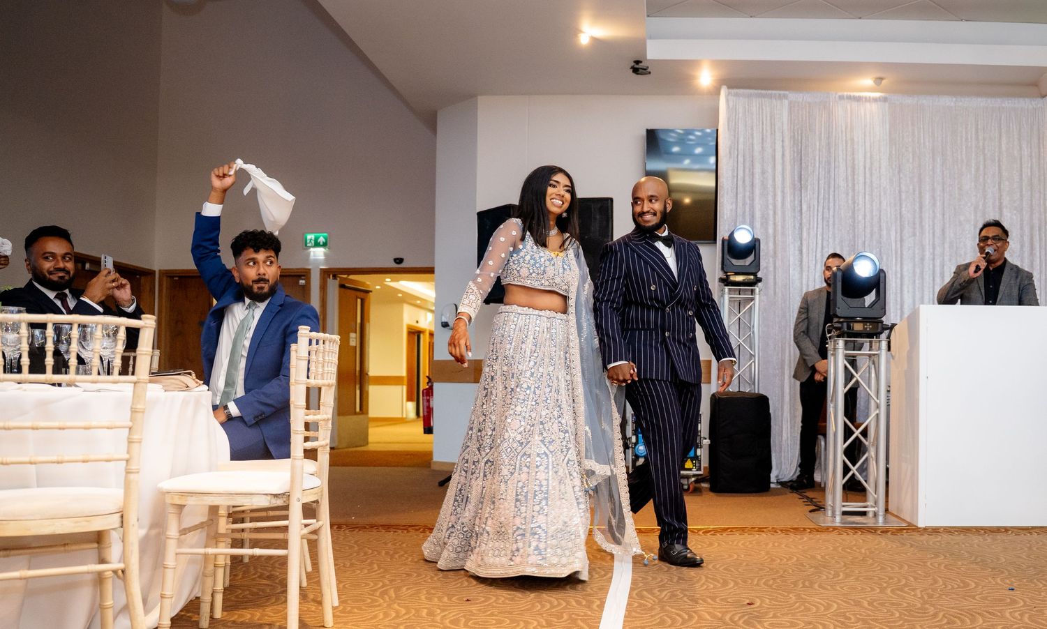 Bride in silver lehenga and groom in striped suit make grand entrance at wedding reception.