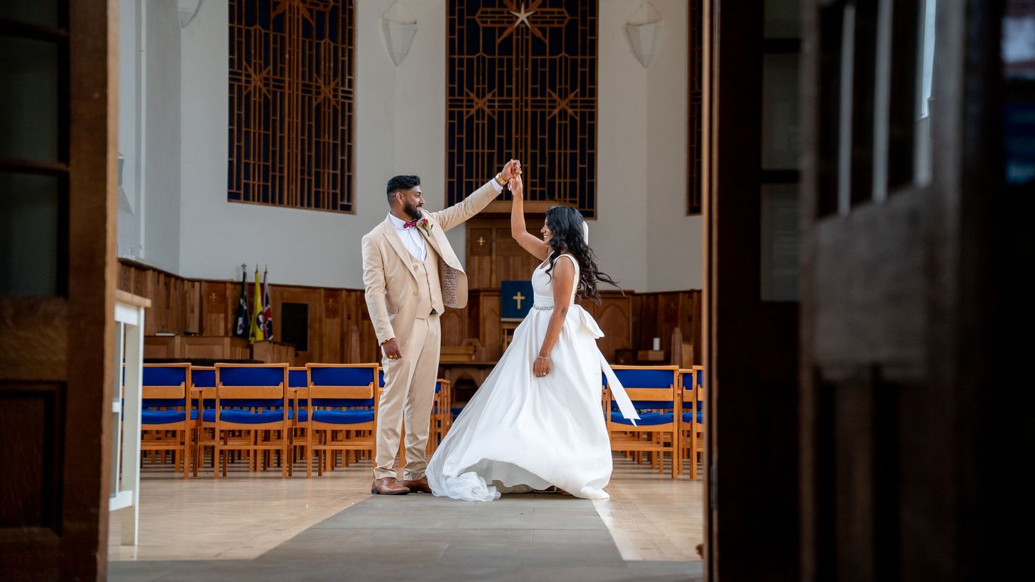 Bride and groom share a romantic dance in a sunlit church aisle, groom in beige suit twirling bride in white gown.