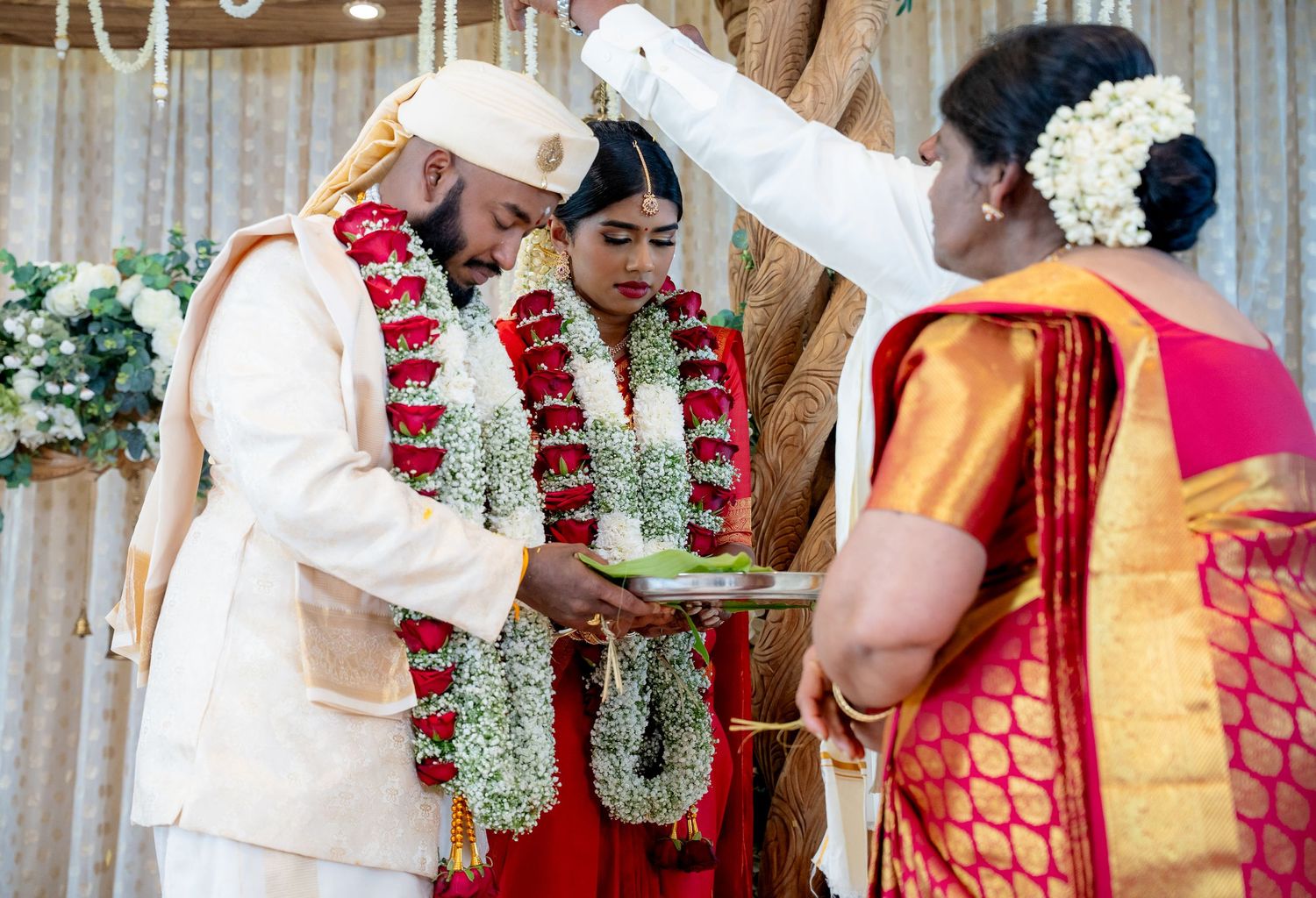 Elder woman performs traditional blessing ritual for bride and groom wearing flower garlands at South Indian wedding.