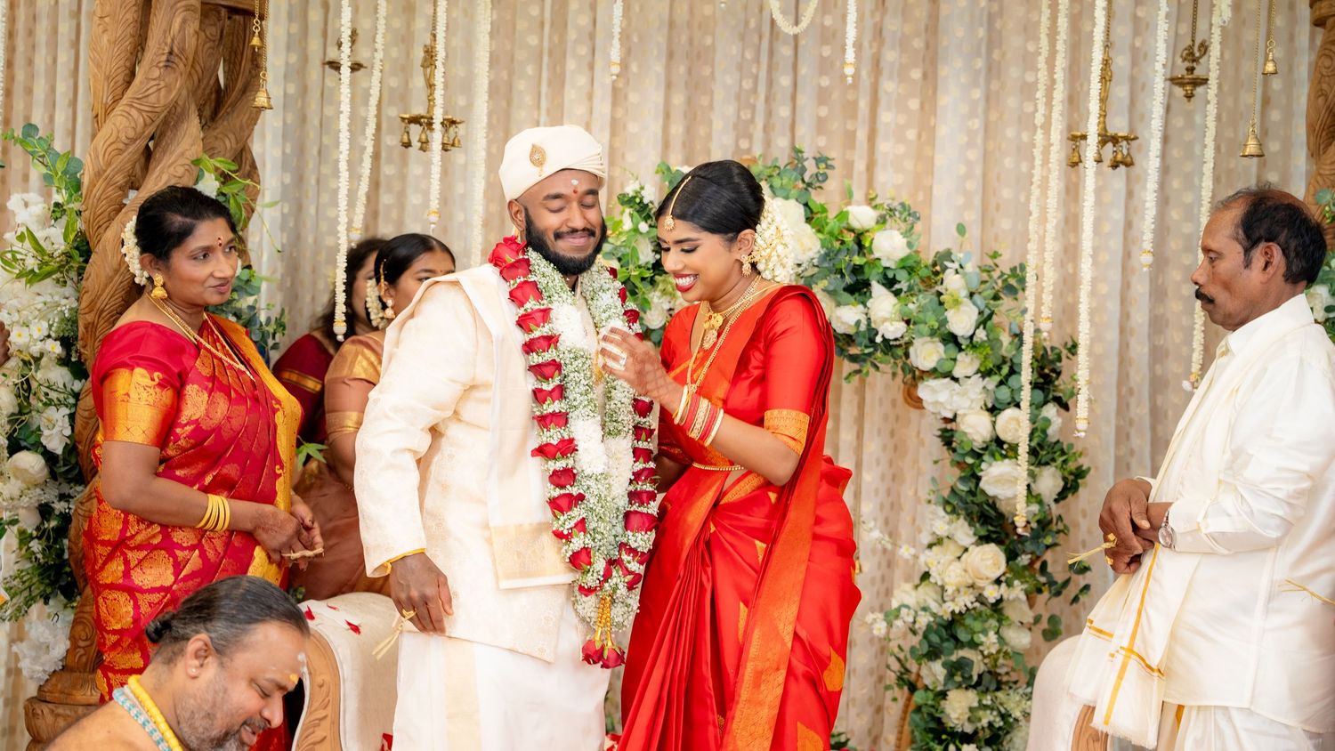 Newlywed couple smiles wearing flower garlands during joyful South Indian Hindu wedding ceremony.