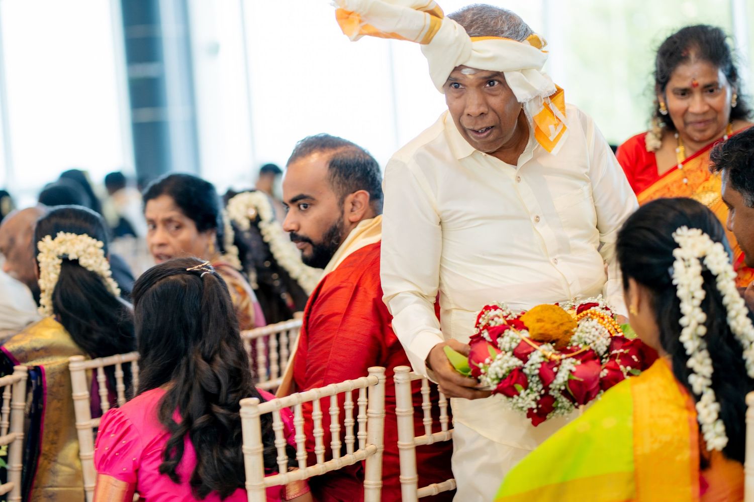 Elder man in white traditional attire carries floral garlands through seated wedding guests at Indian ceremony.