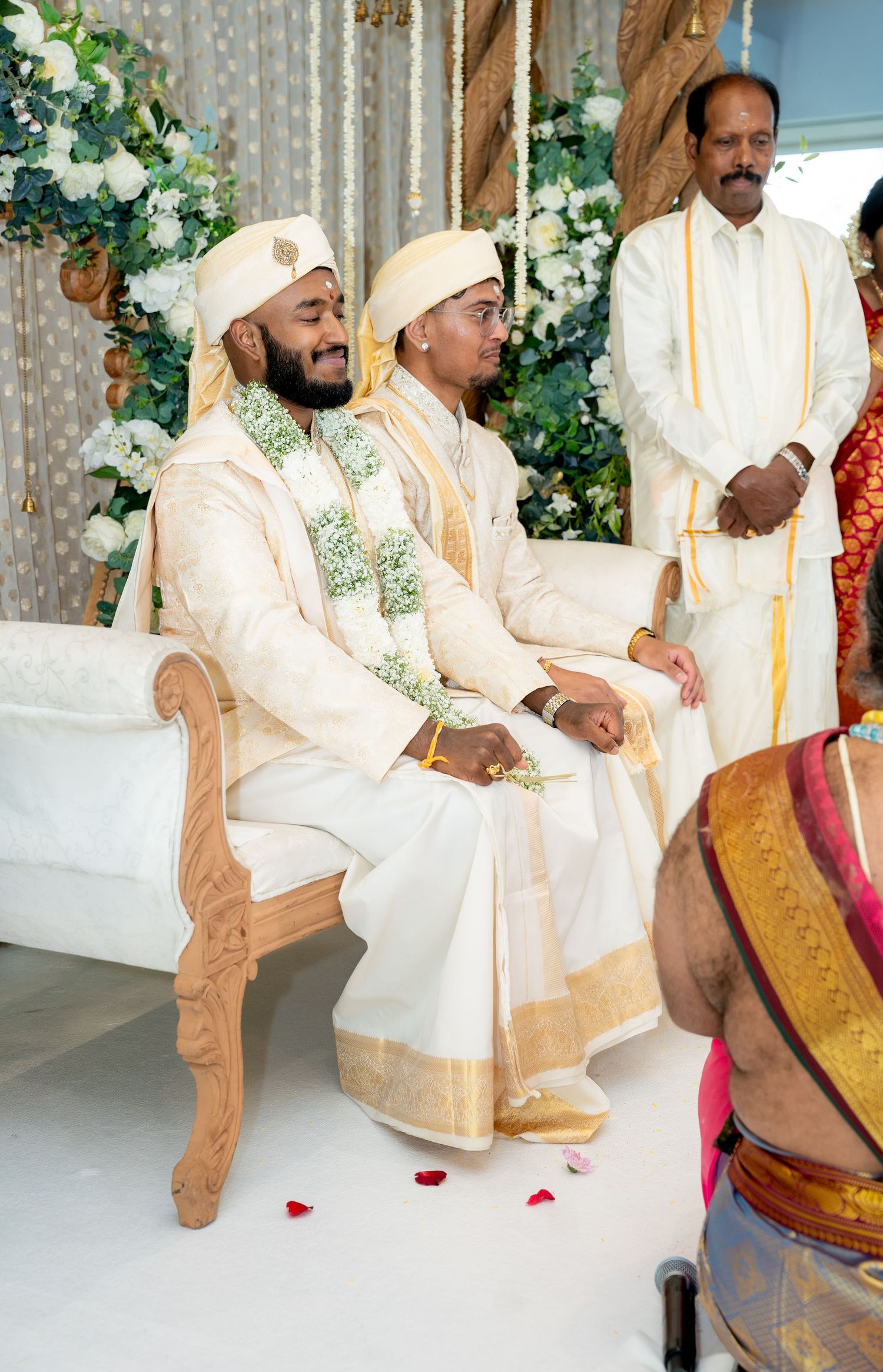 Two grooms in matching cream sherwanis with floral garlands seated at traditional South Indian wedding mandap.