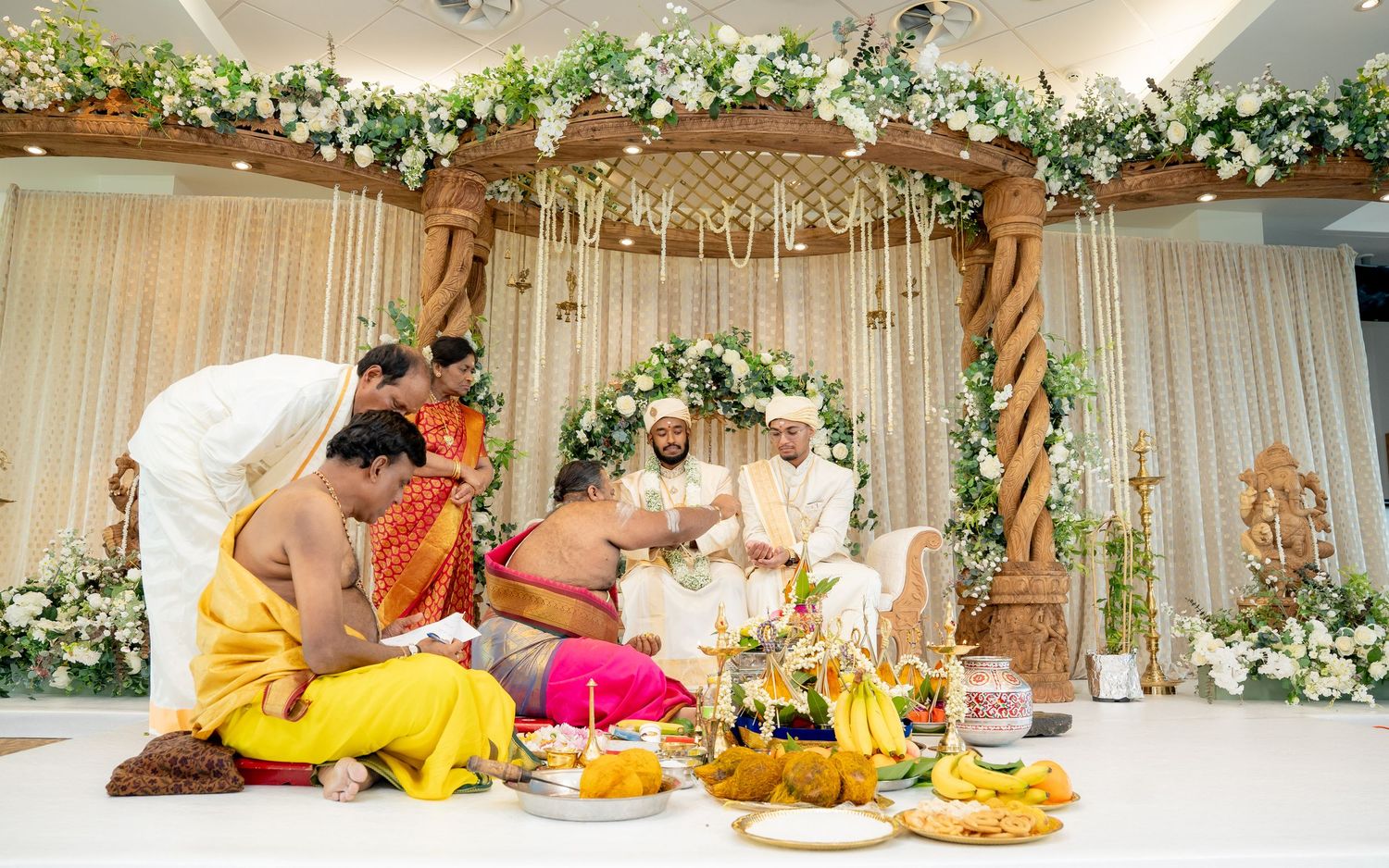 Hindu wedding ceremony with two grooms seated on a decorated mandap with priests performing rituals.