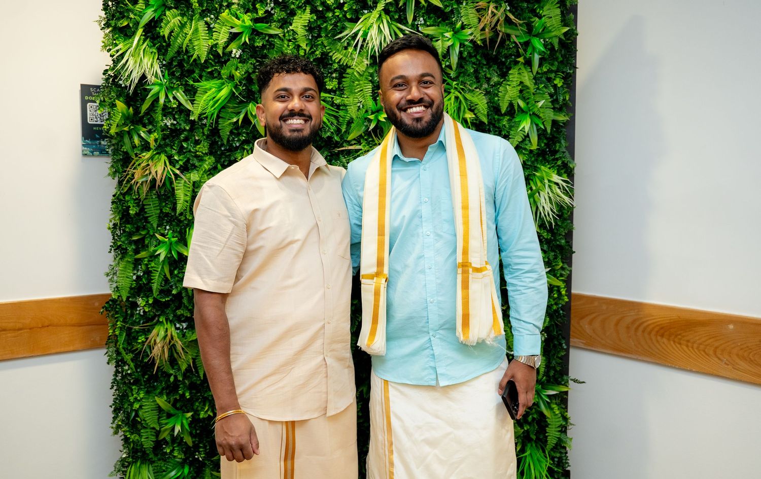 Two men in traditional South Indian white dhotis smiling together in front of a green plant wall.