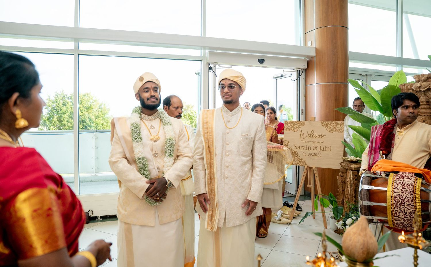 Two men in cream sherwanis and golden turbans standing at a decorated traditional Hindu wedding ceremony venue.
