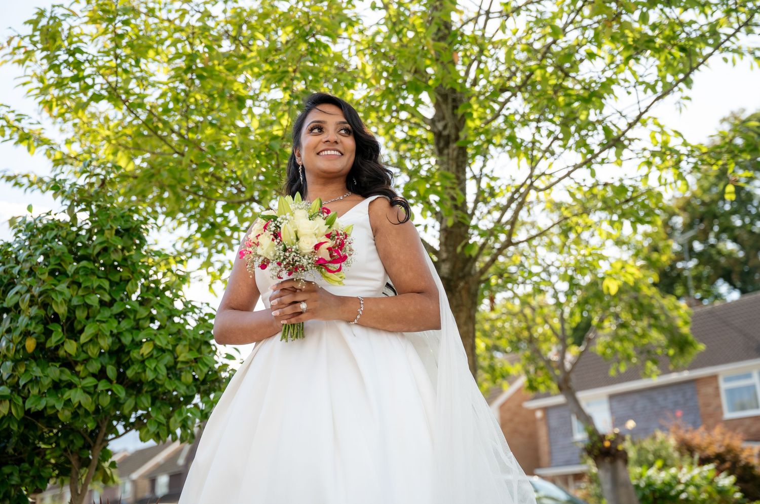 Smiling bride in white ball gown holding colorful bouquet outdoors under green trees on her wedding day.