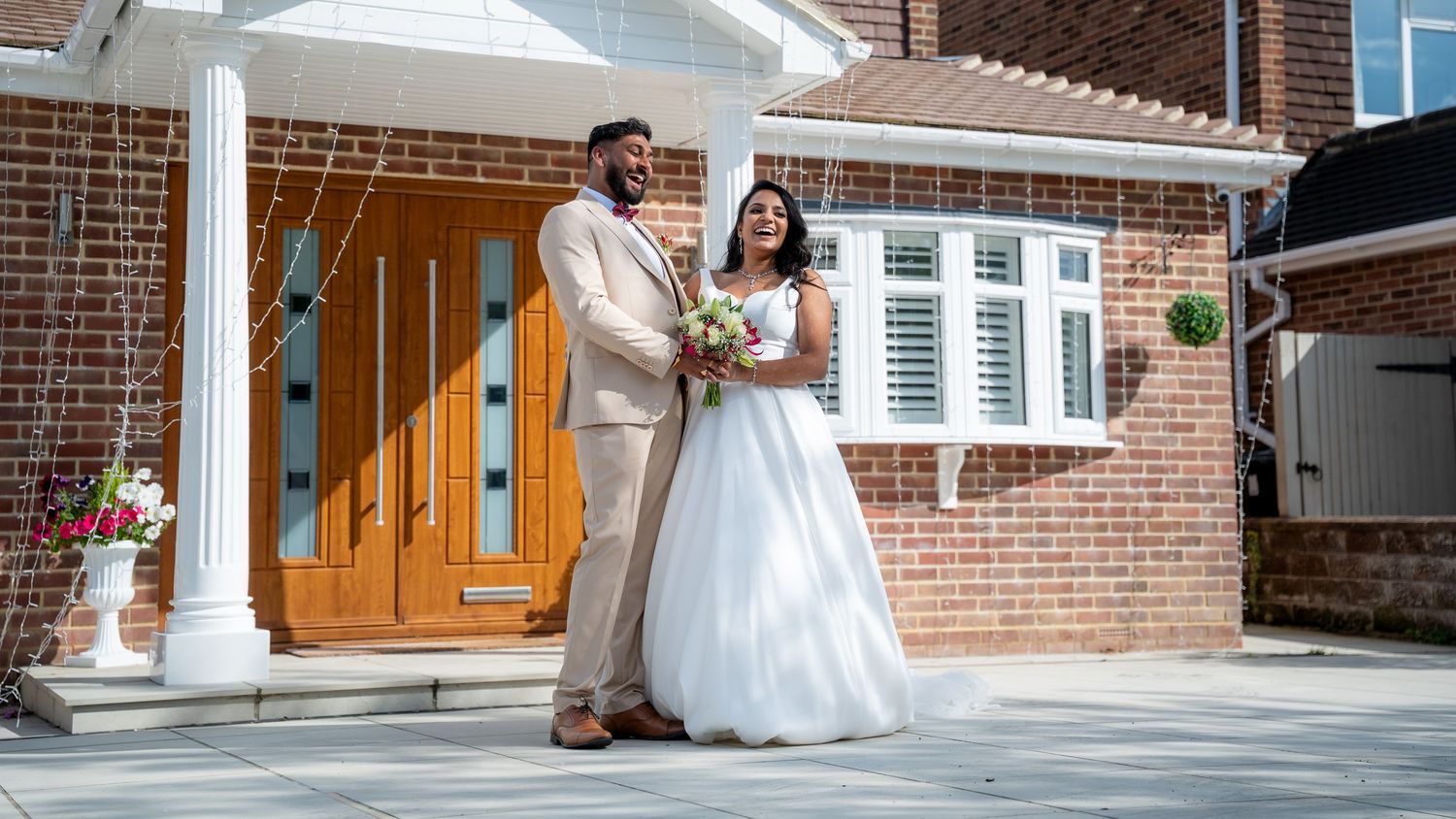 Bride and groom posing in front of a brick house with a wooden door on their wedding day.