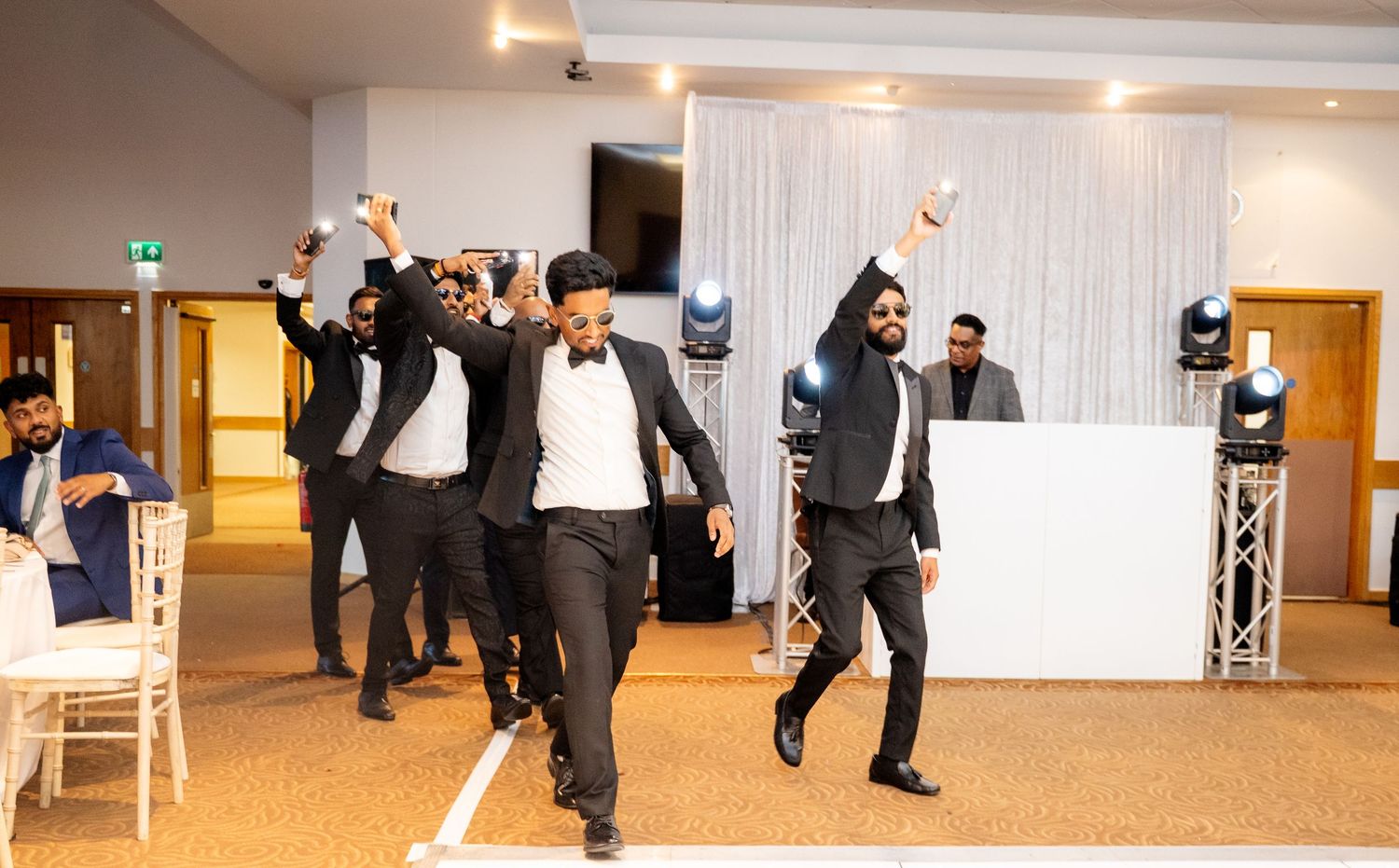 Groomsmen in black tuxedos dancing with raised arms at a wedding reception with DJ setup in background.
