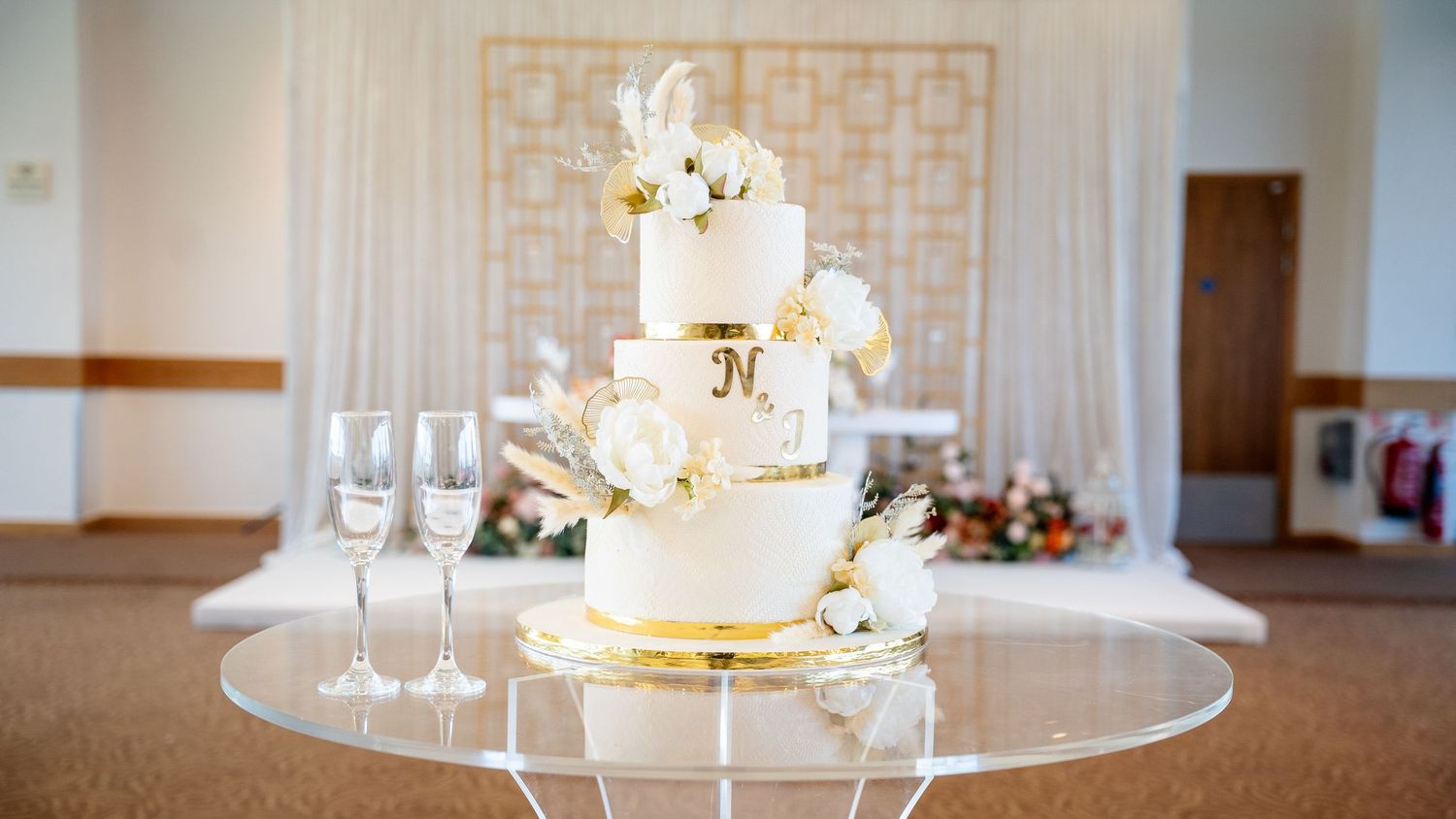 Three-tier white wedding cake with gold accents and floral decorations on glass table in wedding hall.