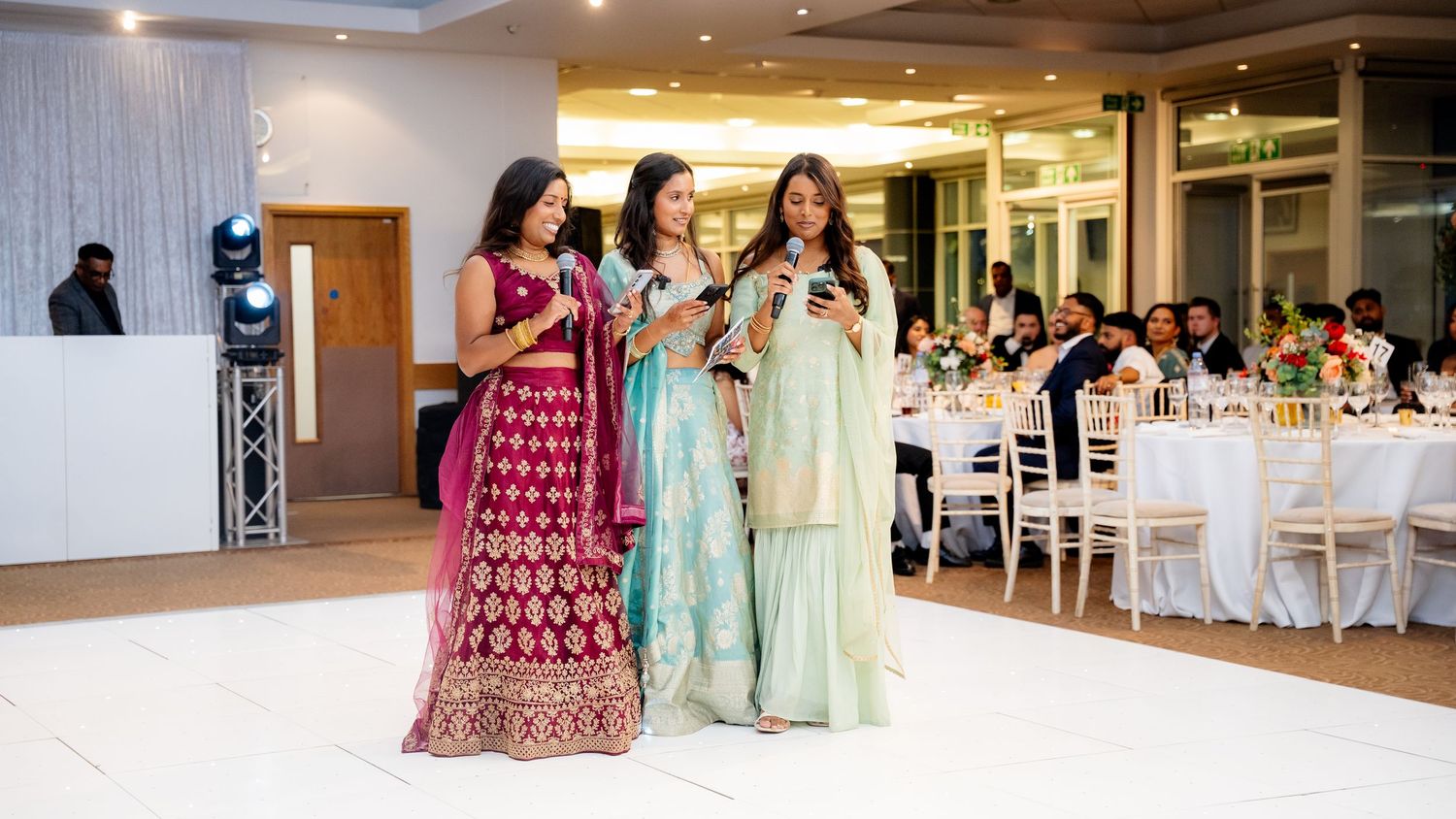 Three women in colorful Indian outfits perform with microphones on a white dance floor at a wedding.