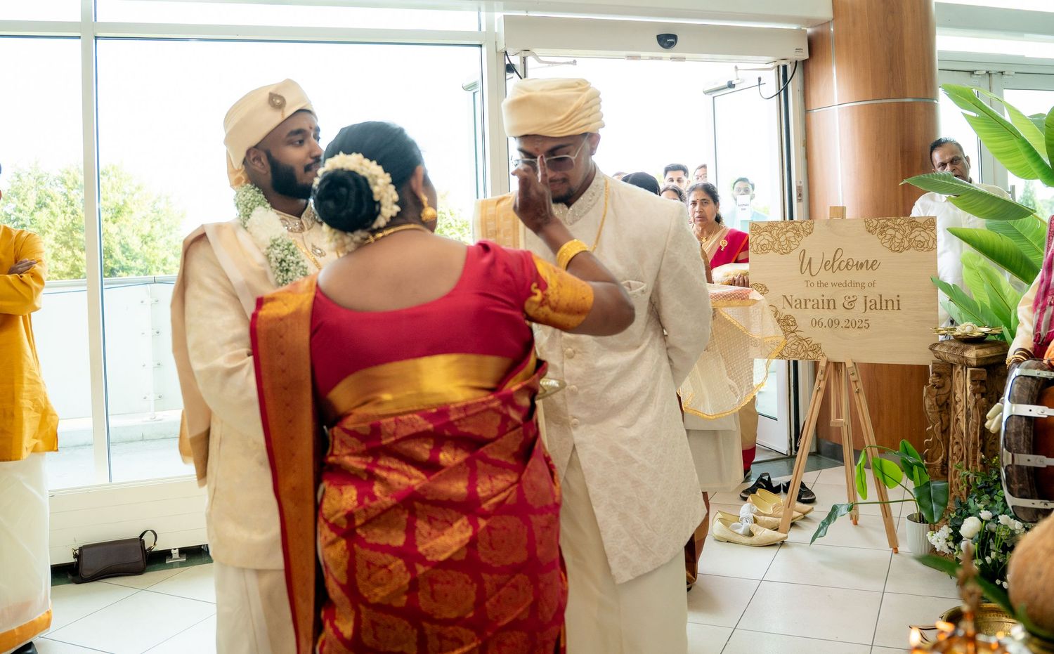 Groom in cream sherwani greeting a bride in red and gold saree at a traditional Hindu wedding ceremony entrance.