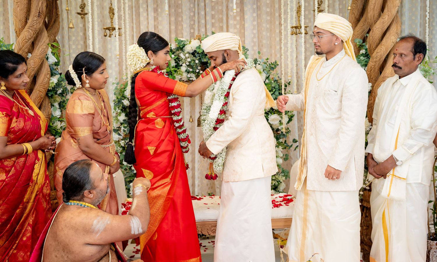 South Indian bride and groom perform Jaimala garland ceremony with priest and family members present.