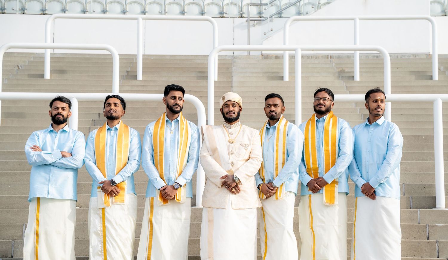 Seven men in light blue kurtas with gold sashes flanking groom in white on stadium steps.