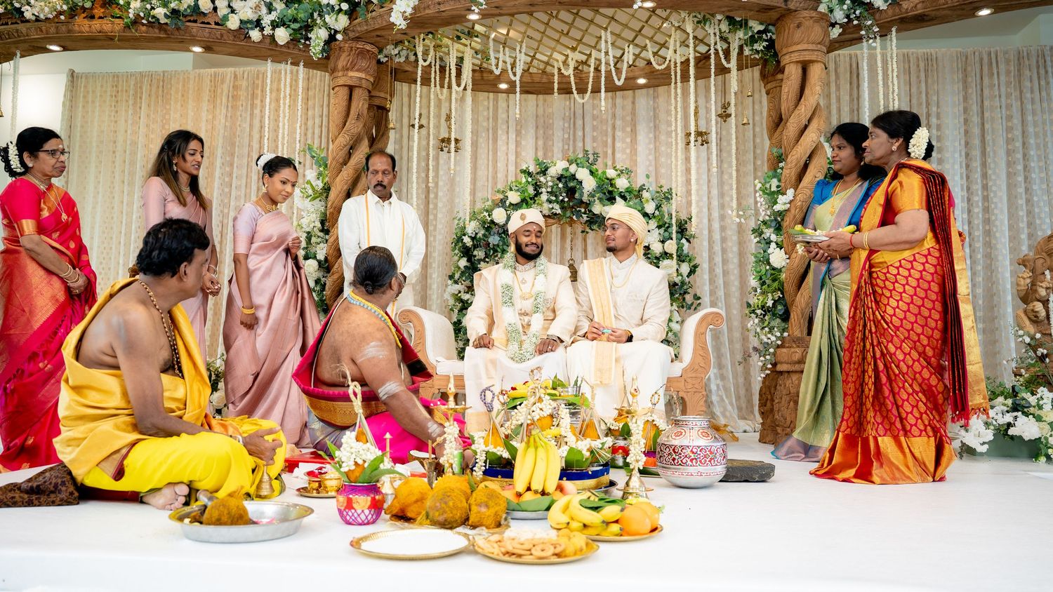 Traditional South Indian wedding ceremony with guests in colorful sarees gathered around a decorated mandap with floral arrangements.