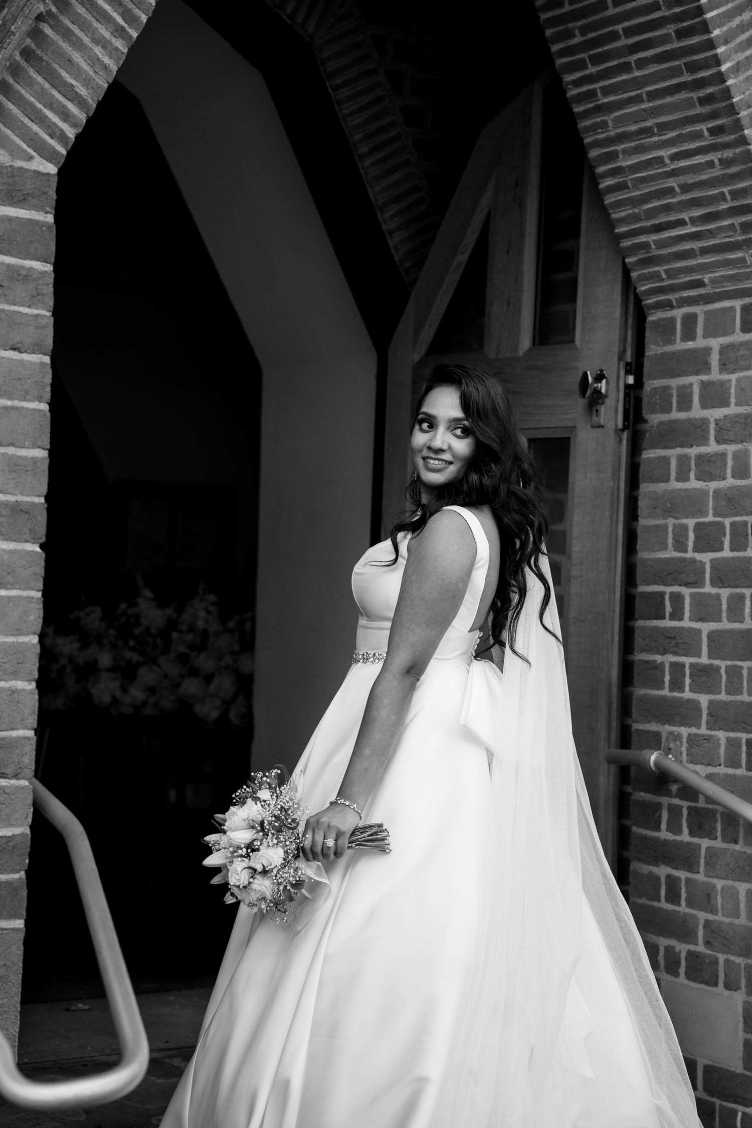 Black and white photo of a smiling bride in a white gown holding a bouquet, posing by a brick archway.