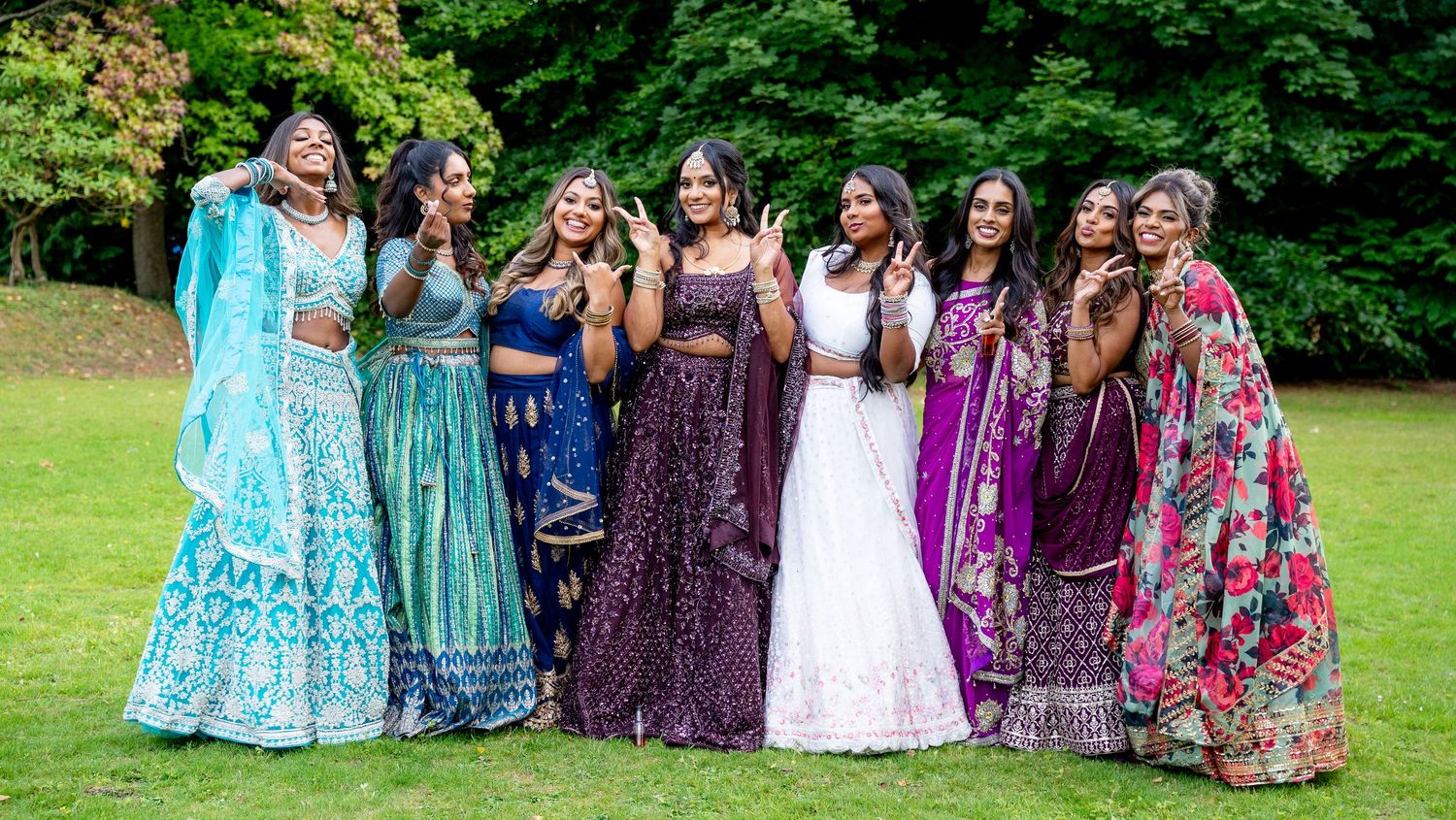 A group of women in colorful Indian lehengas smile and pose together on a green lawn.