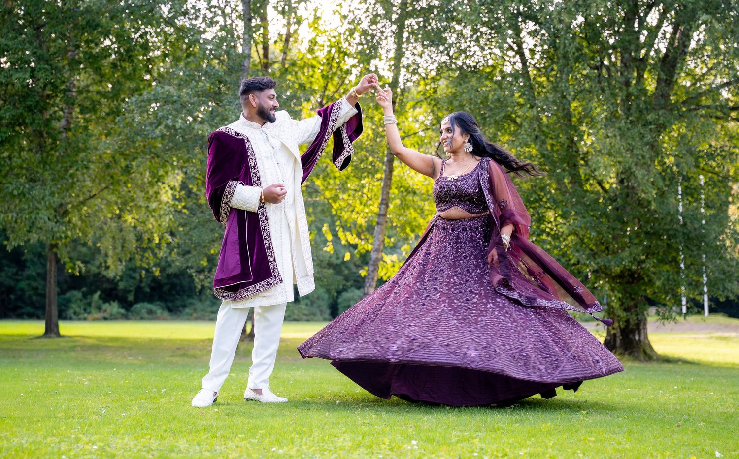 Indian wedding couple dancing on grass, bride in sparkling purple lehenga, groom in white and purple sherwani.