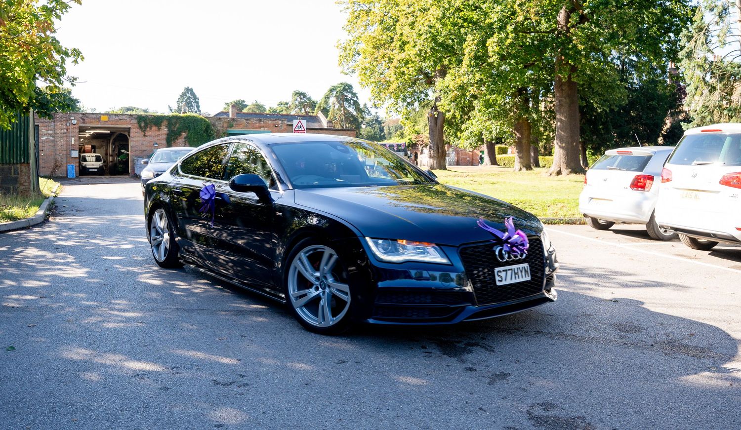 Black Audi A7 wedding car decorated with purple ribbon and bow parked outdoors.