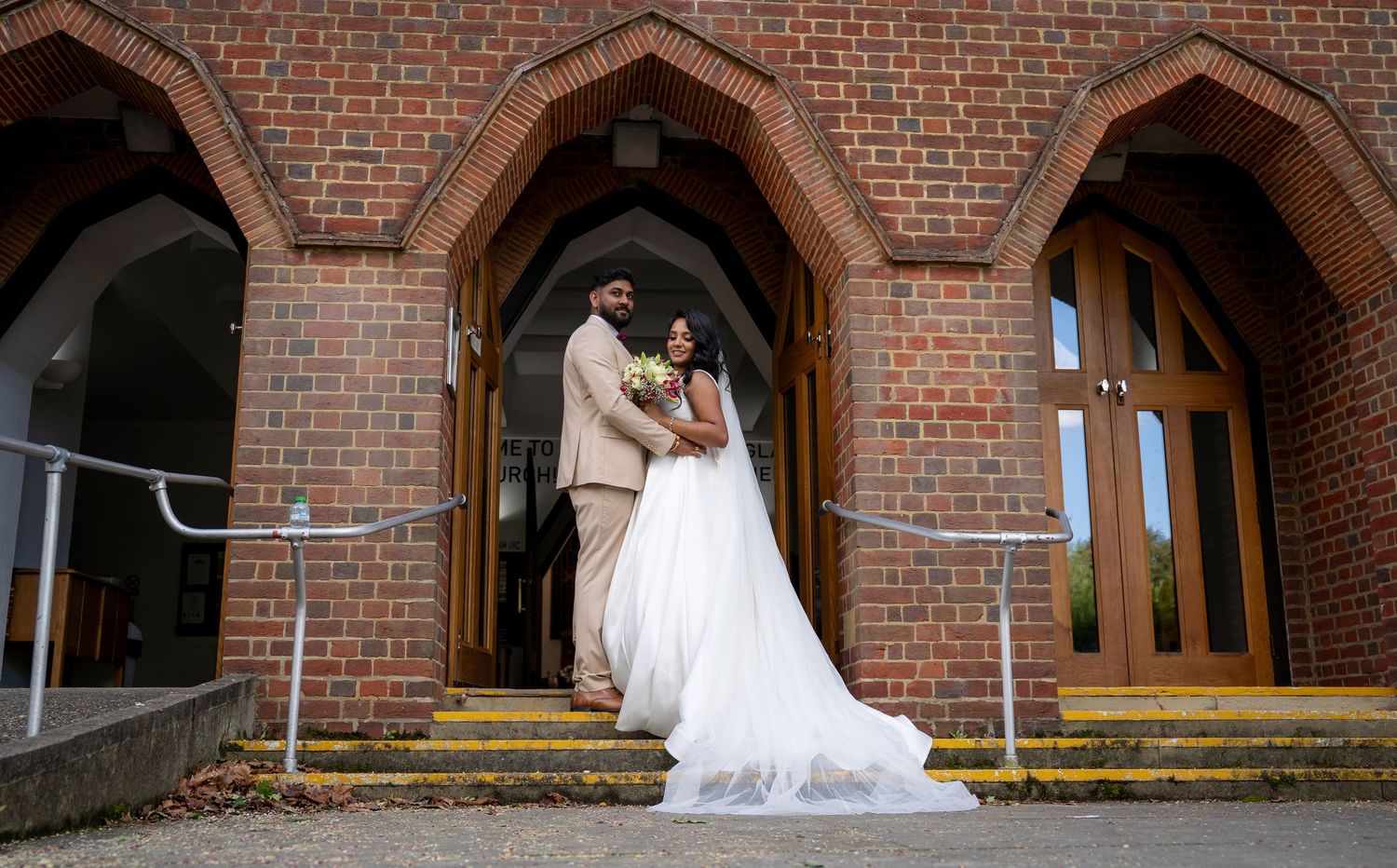 Bride and groom posing at brick church entrance, bride in white gown, groom in beige suit, wedding photography.