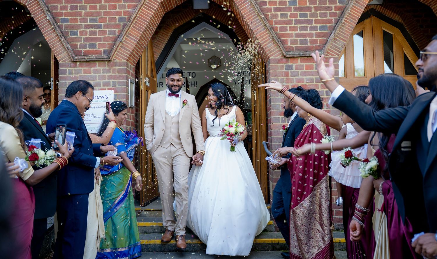 Bride and groom exit church amid confetti celebration, guests in colorful traditional attire surround them.