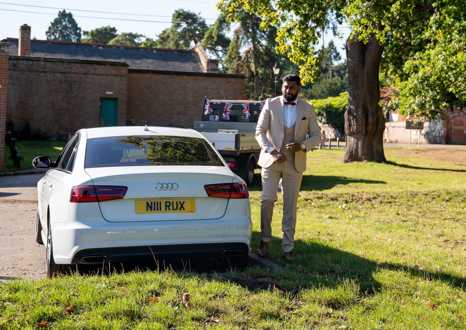 Man in beige suit stands beside white Audi with personalized number plate N111 RUX on green lawn.