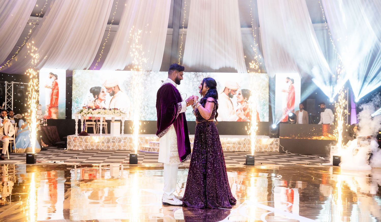 South Asian newlyweds sharing a first dance on a shimmering dance floor with draped fabric backdrop.