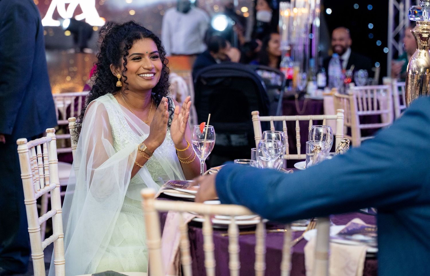 Woman in elegant mint green saree clapping and smiling at a beautifully decorated wedding reception.