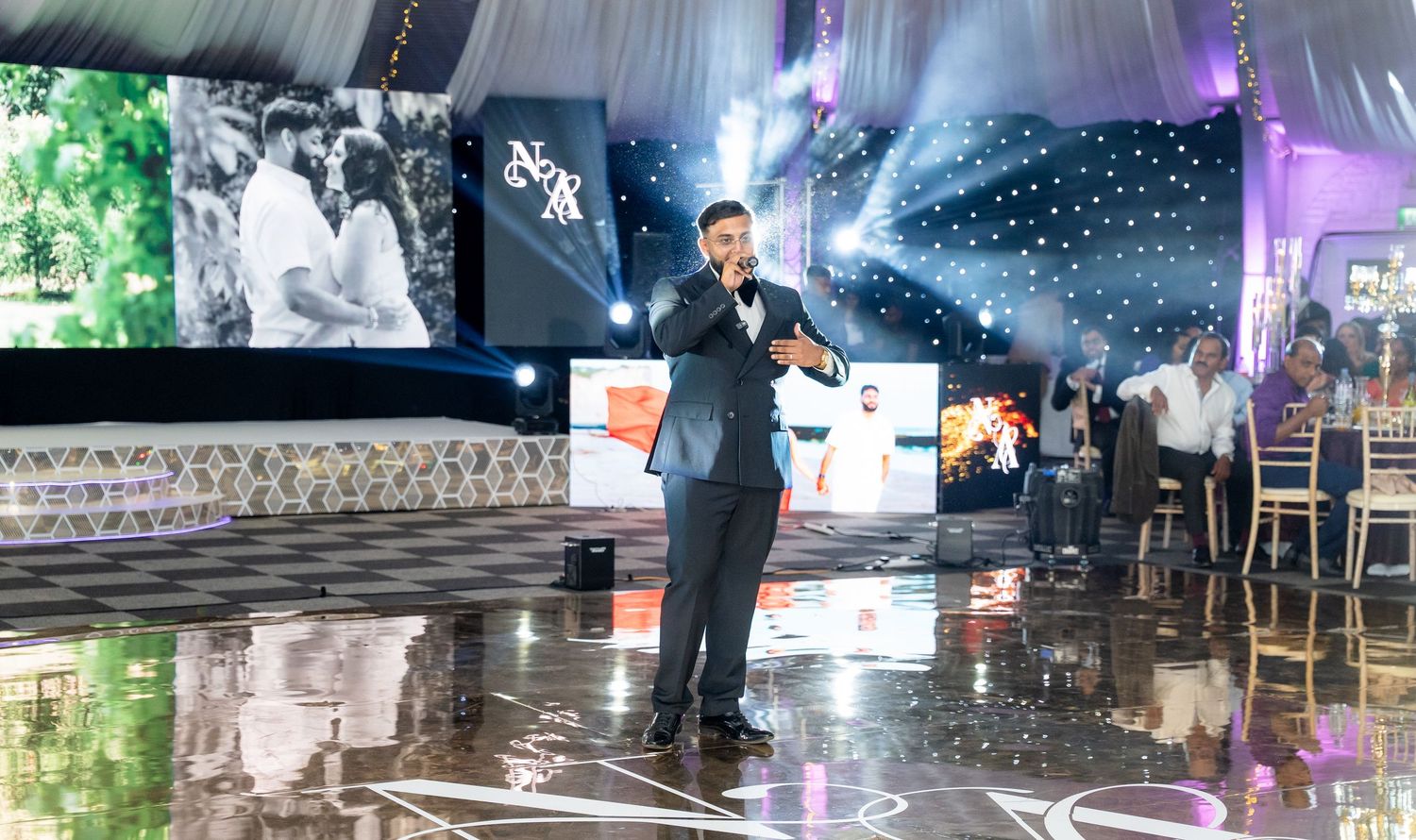 Man in suit performing on stage at elegant wedding reception with LED screens and decorative lighting.