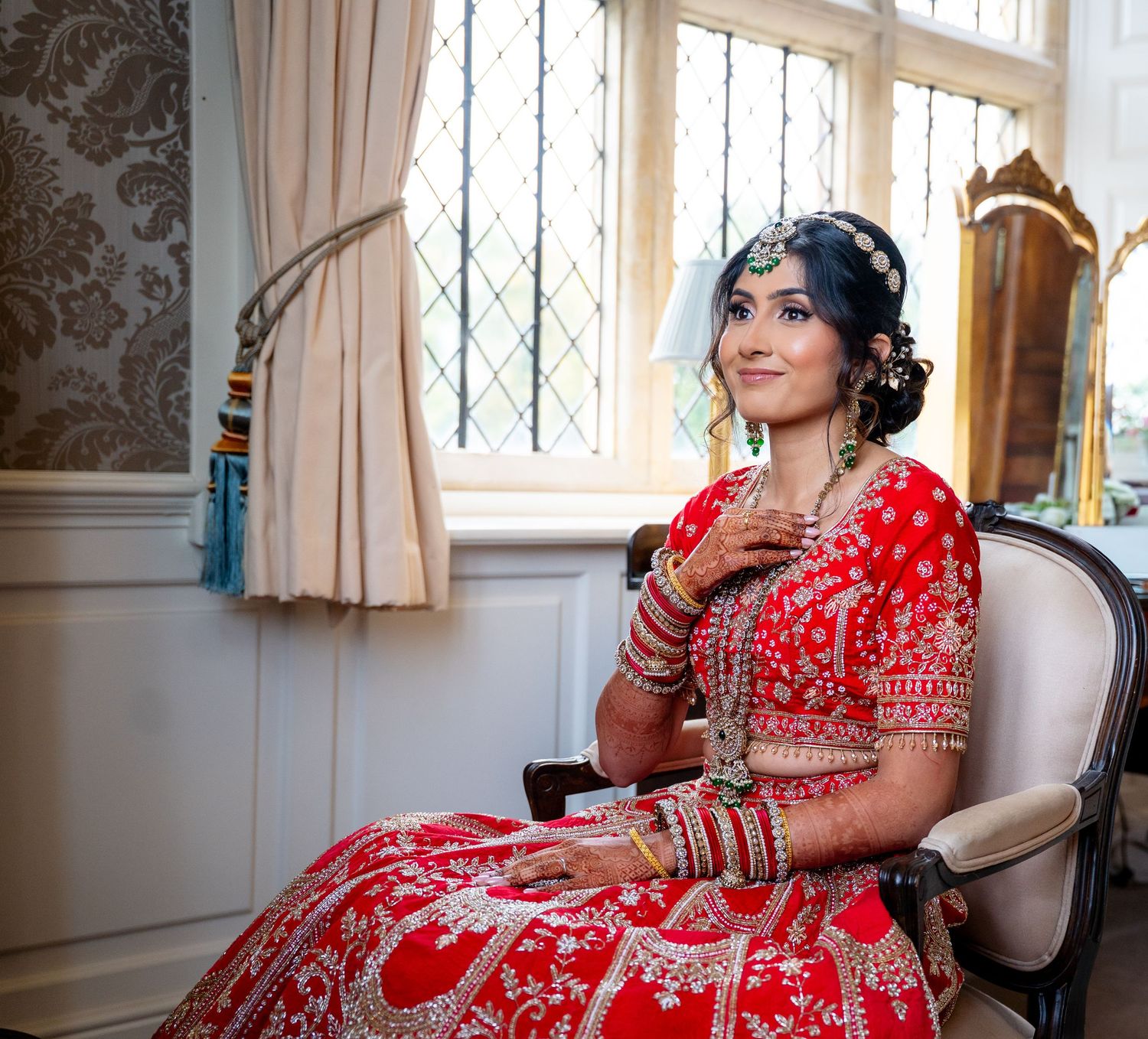 Indian bride in red lehenga and jewelry sits gracefully by window in dressing room.
