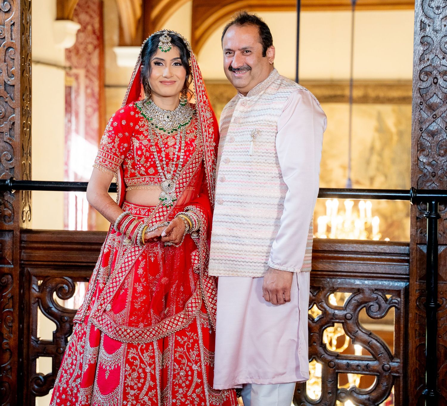 Indian bride in red bridal lehenga posing with father in white kurta at ornate wedding venue.
