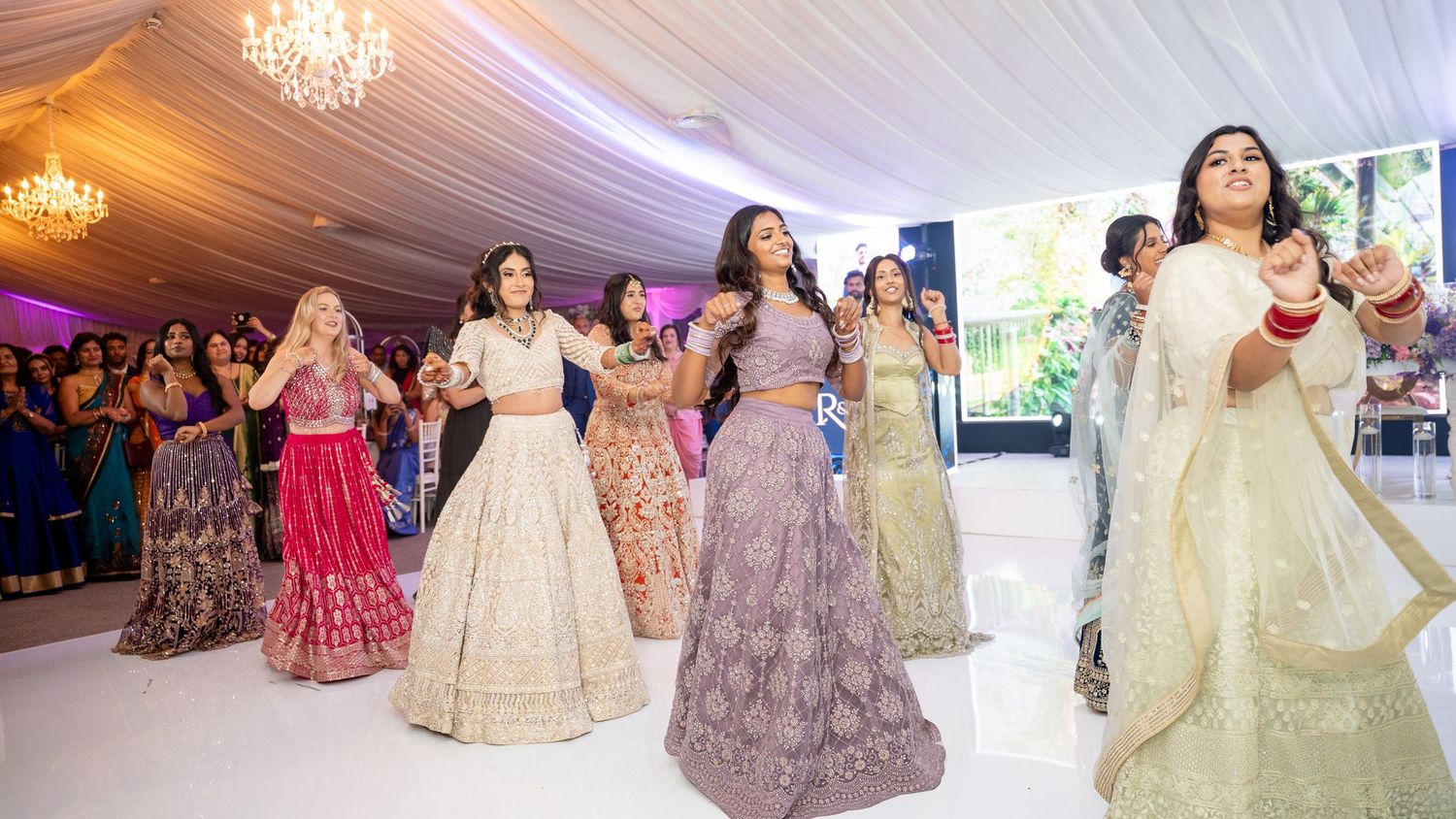 Bridesmaids in colorful Indian outfits perform an energetic dance at a wedding reception.