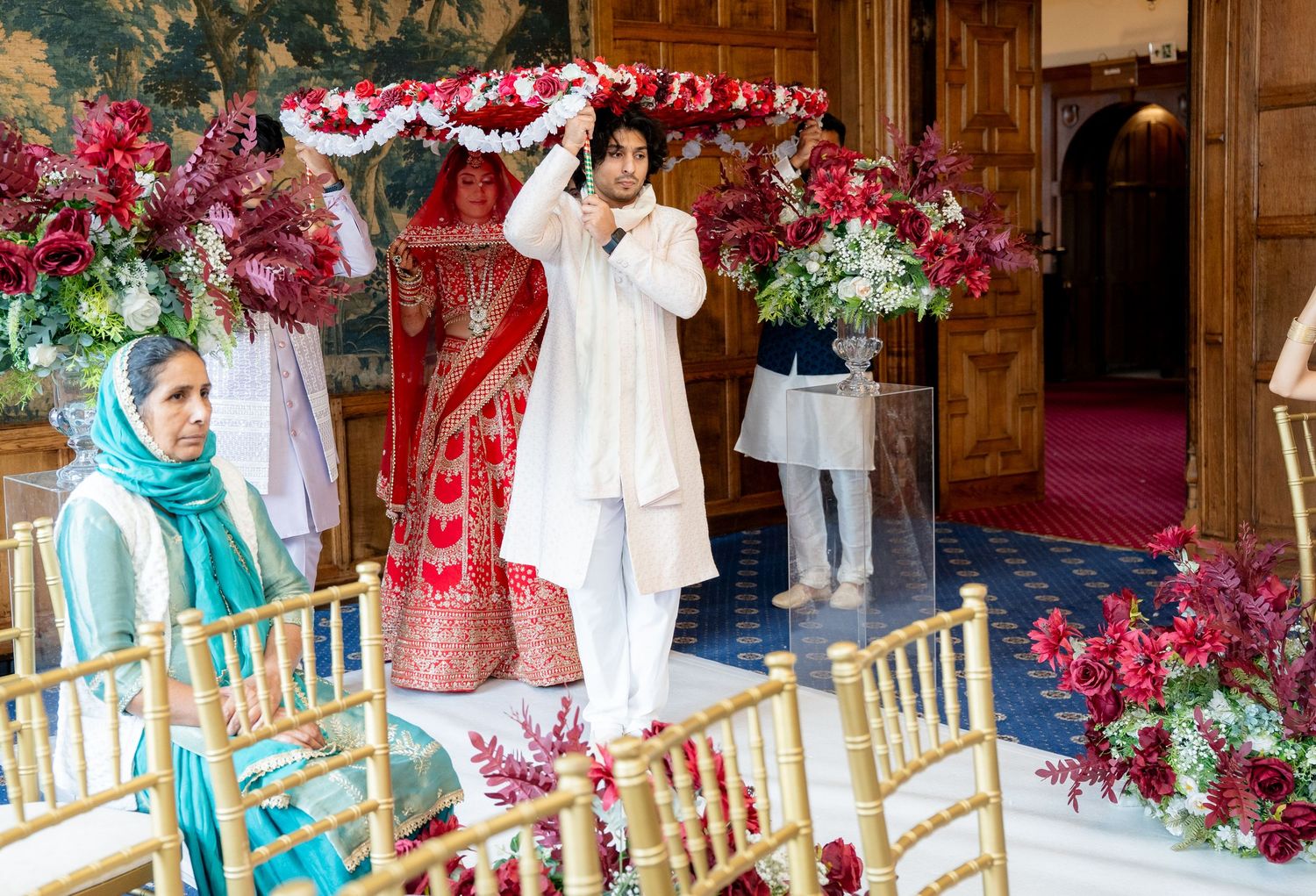 Family holds flower canopy over Indian bride in red lehenga during traditional wedding ceremony.