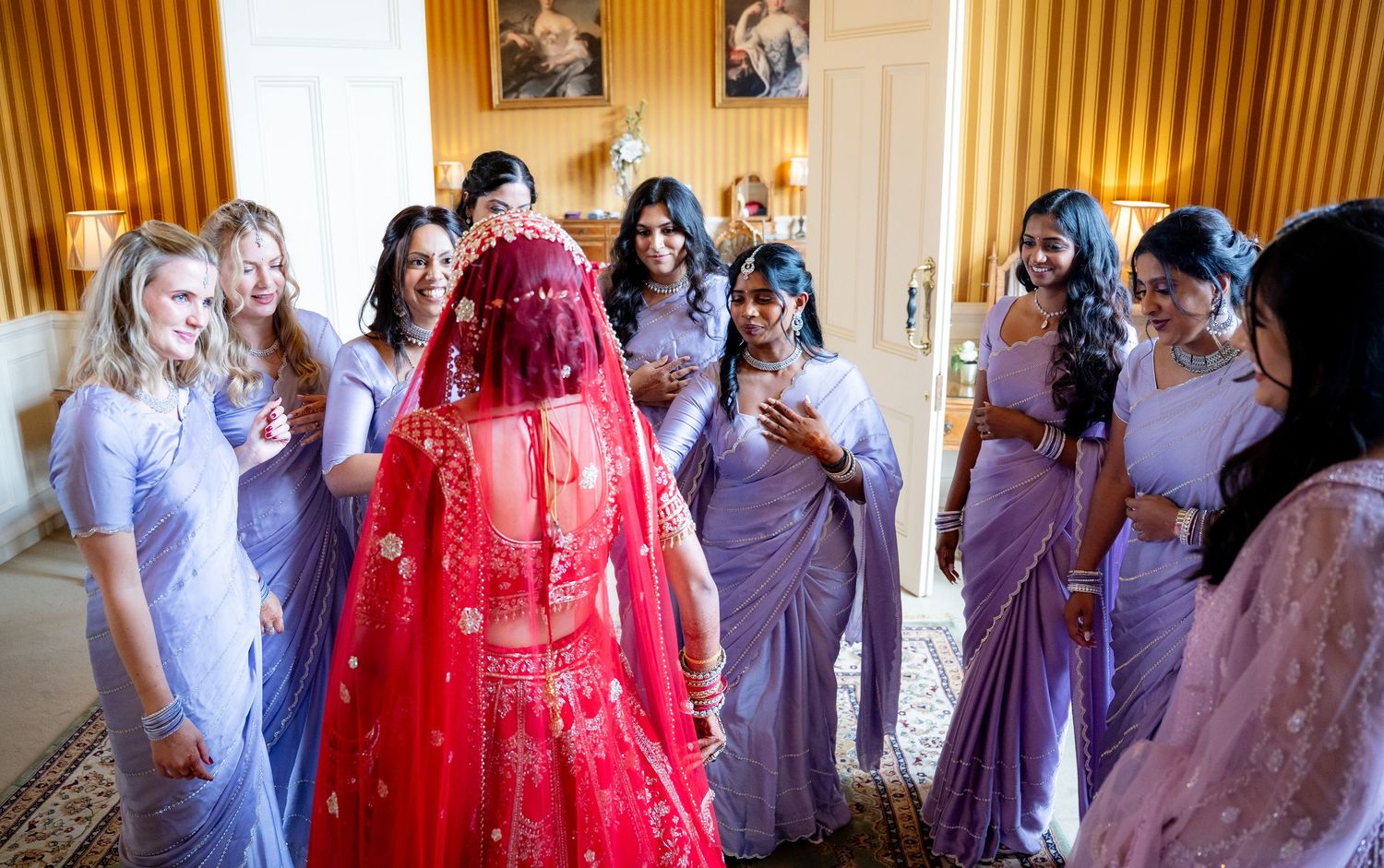 Indian bride in red lehenga revealed to bridesmaids in lavender sarees during bridal preparation.