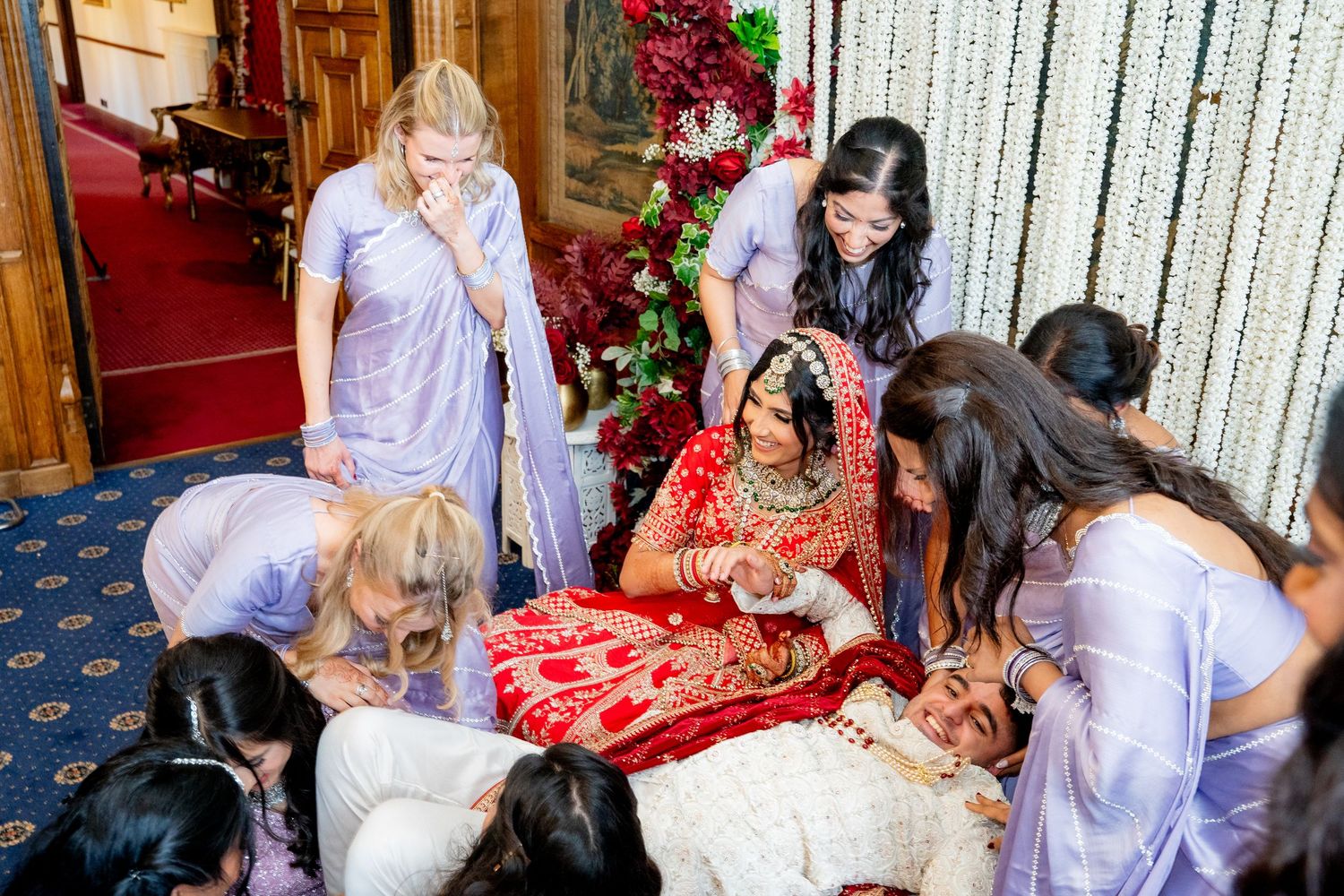 Indian bride in red lehenga surrounded by bridesmaids in lavender during wedding ceremony at ornate venue.