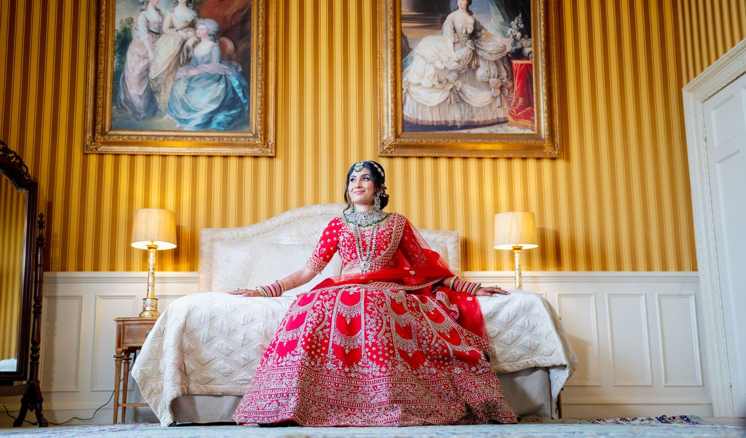 Bride in red embroidered lehenga seated on bed in elegant room with portrait paintings.