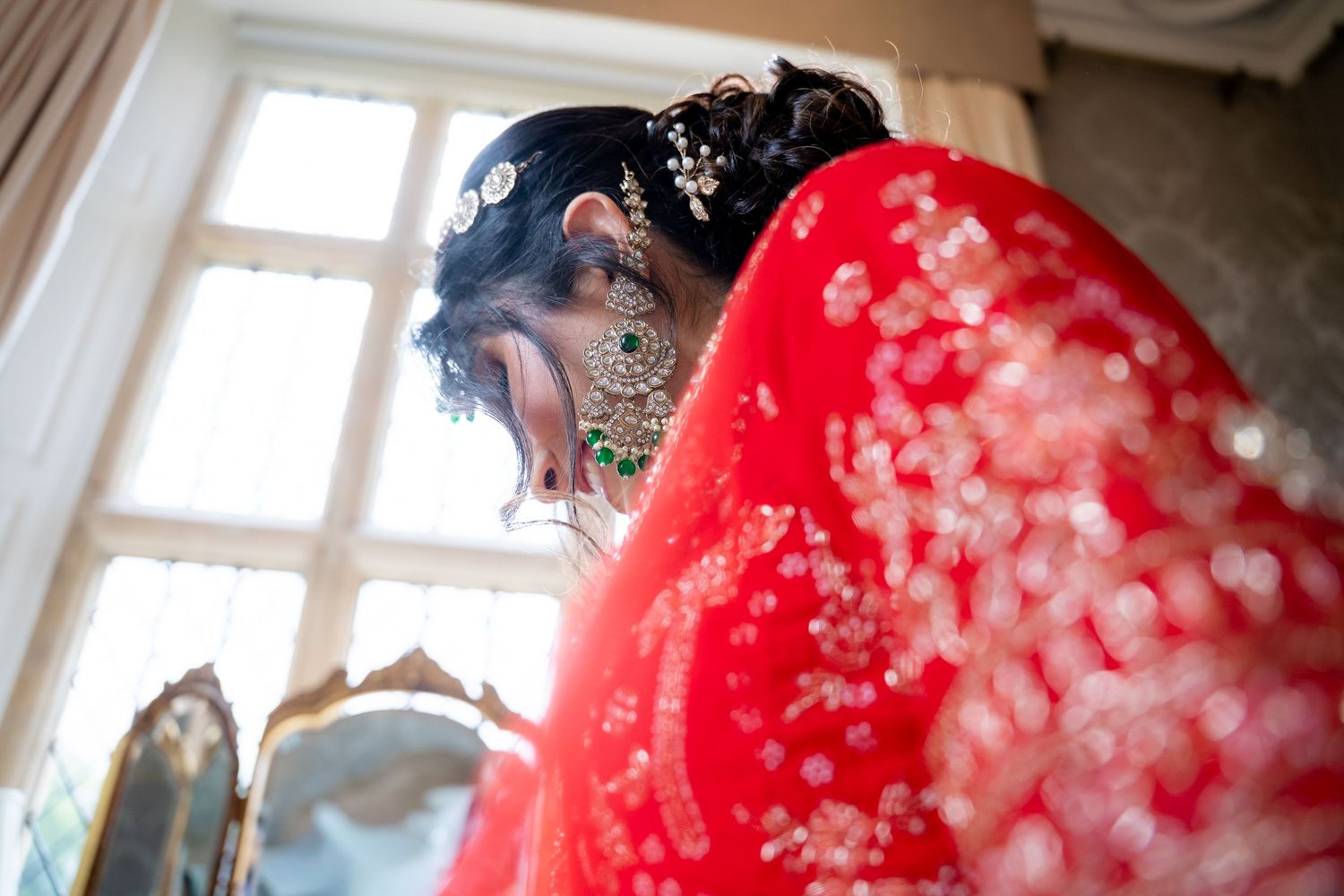 Indian bride in red dupatta with bokeh light effect during bridal preparation near window.
