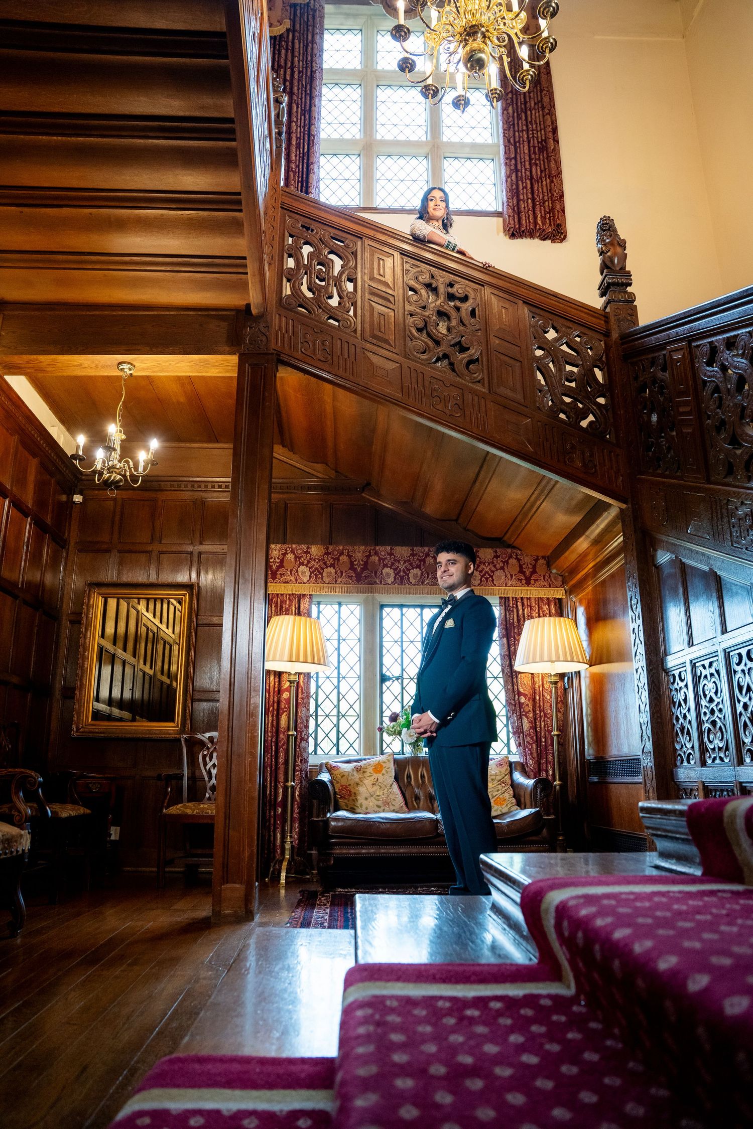 Groom in navy suit stands in ornate wood-paneled hall as bride watches from carved balcony above.