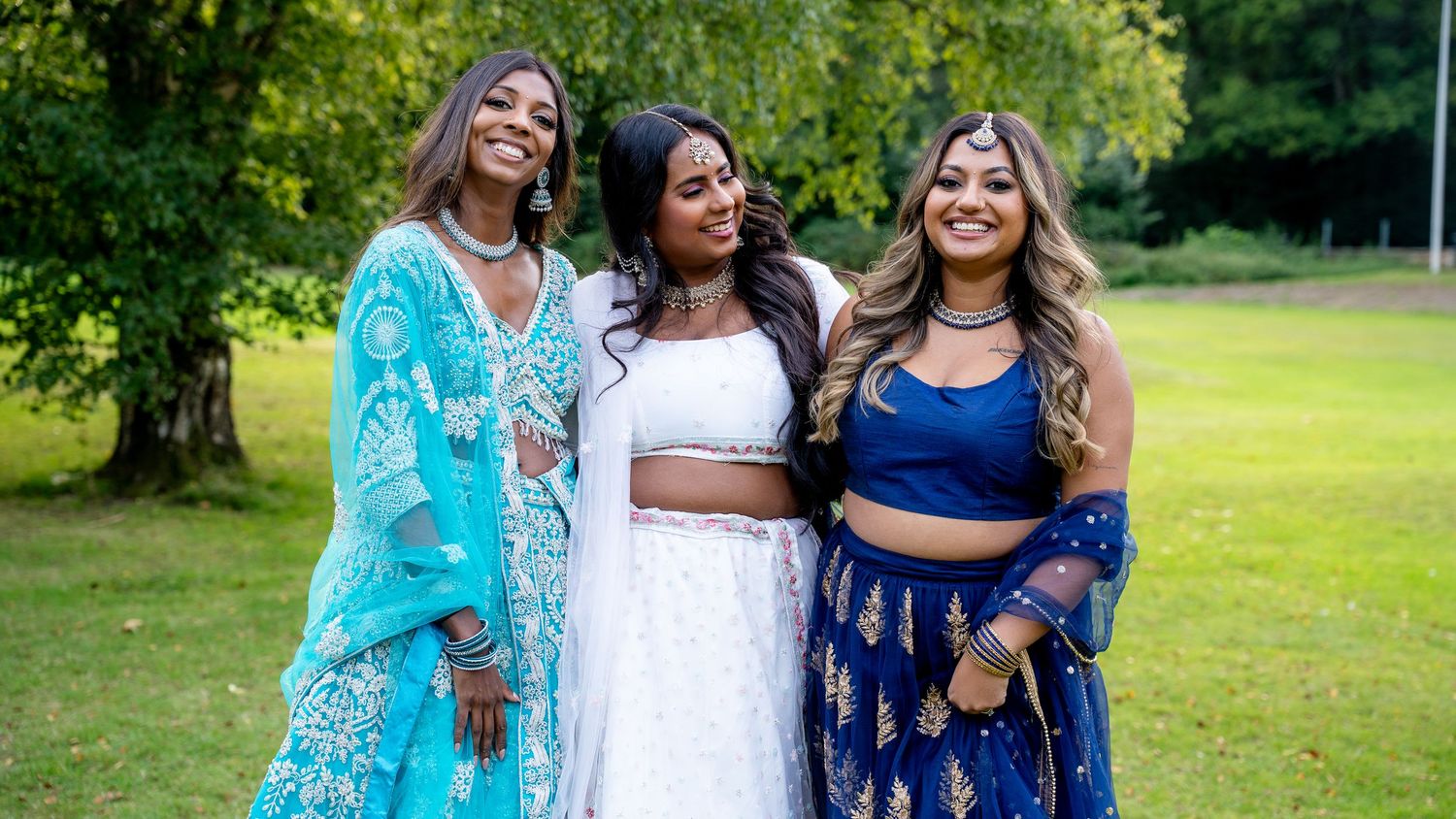 Three women in elegant Indian attire smile together outdoors at a garden celebration.