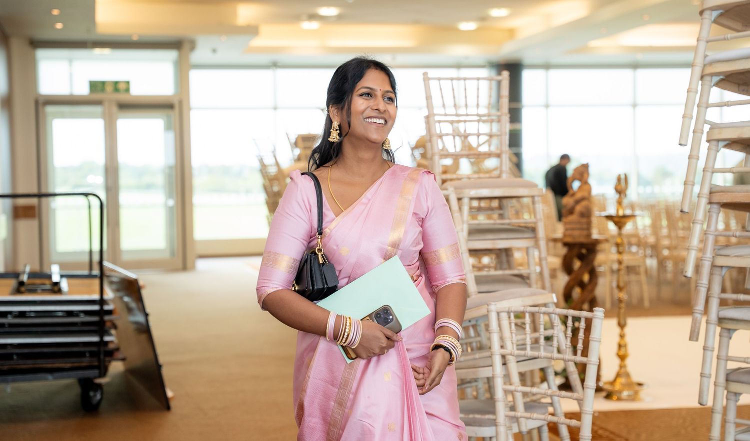 Elegant woman in pink saree carrying a black handbag and blue folder at an indoor wedding venue.