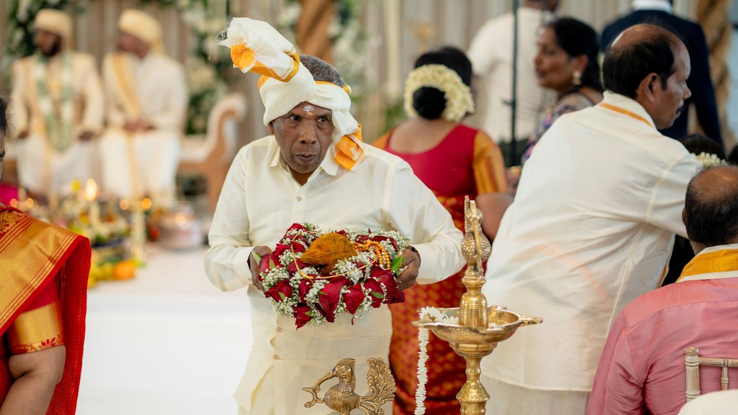 Elder man in white dhoti and turban carries floral offerings during traditional South Indian wedding ritual.