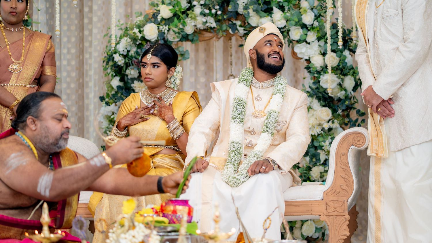 A bride and groom in traditional South Asian wedding attire sit during a Hindu ceremony with a priest and floral decorations.