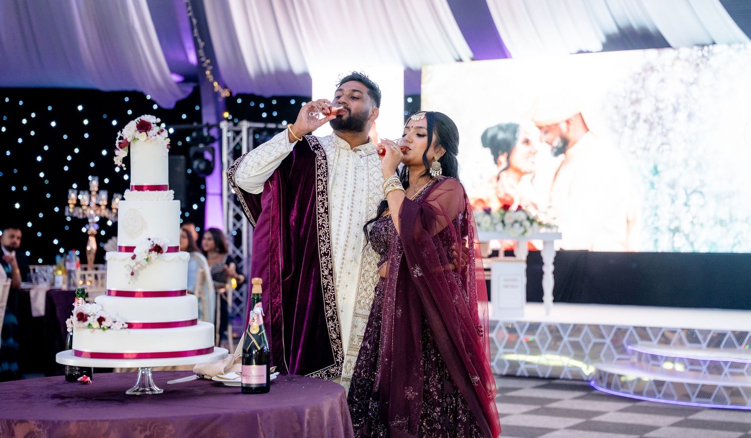 Groom in velvet sherwani drinking champagne with bride in burgundy lehenga at South Asian wedding reception.