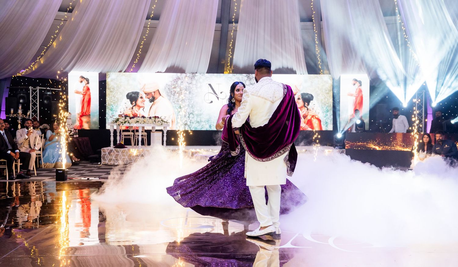 Couple dancing at Indian wedding reception with purple lehenga, fog effects, and vibrant stage backdrop.
