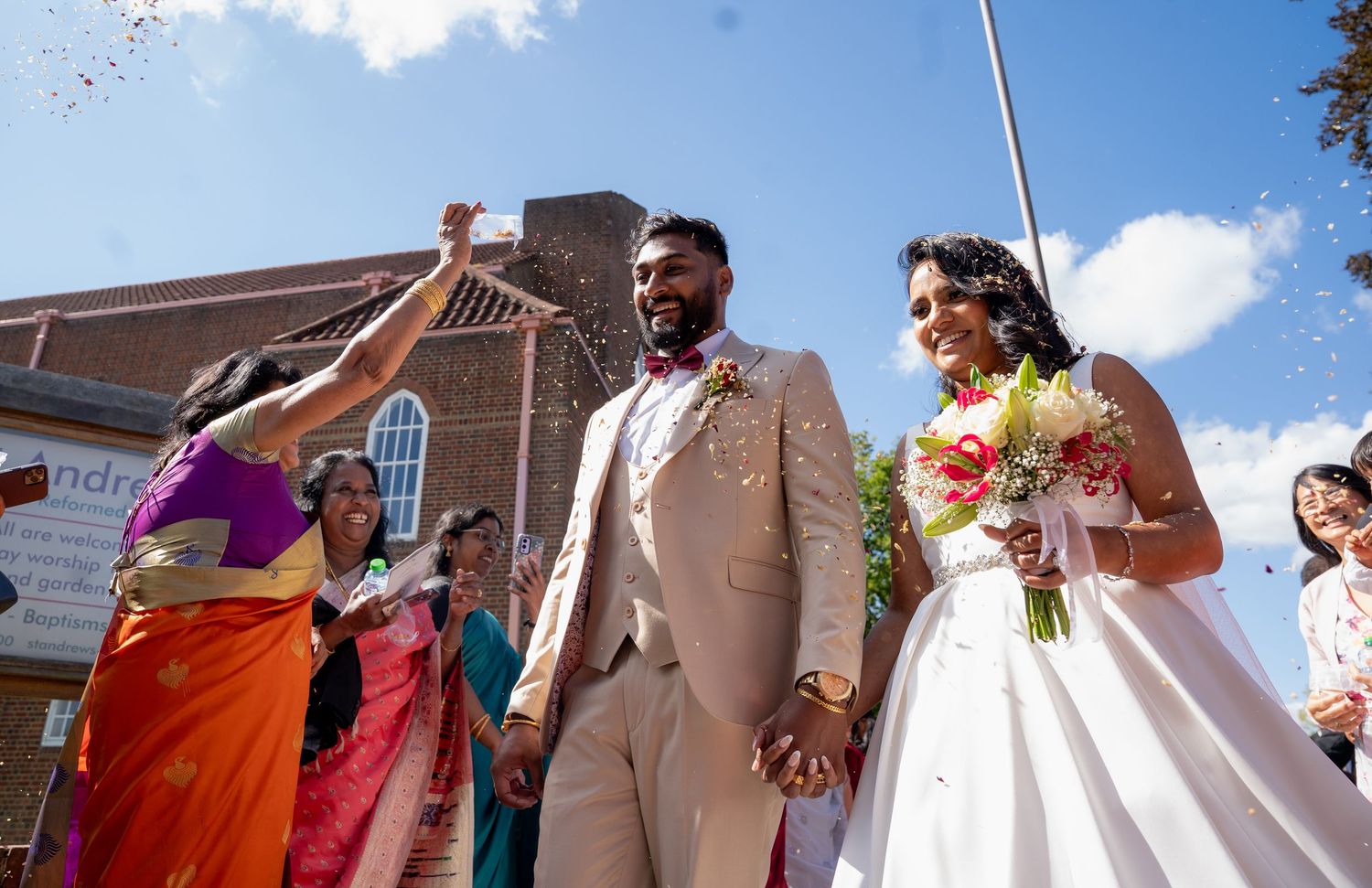 Bride and groom celebrate outside a church as guests shower them with confetti on a sunny day.