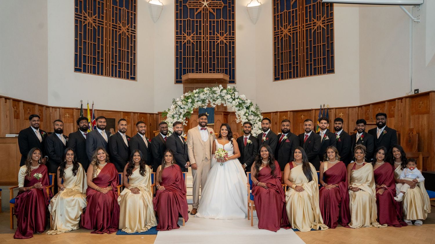 Wedding party poses in church with bride and groom centered, bridesmaids in red sarees and groomsmen in dark suits.