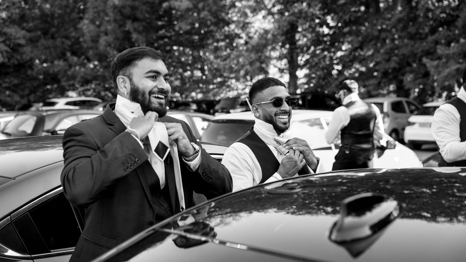 Two groomsmen in suits laugh and celebrate together beside a car in black and white photo.