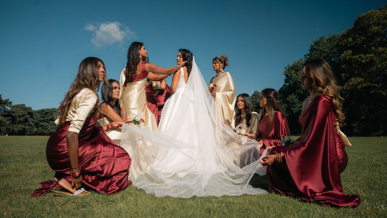 Bride in white gown surrounded by bridesmaids in burgundy and gold dresses on green lawn under blue sky.